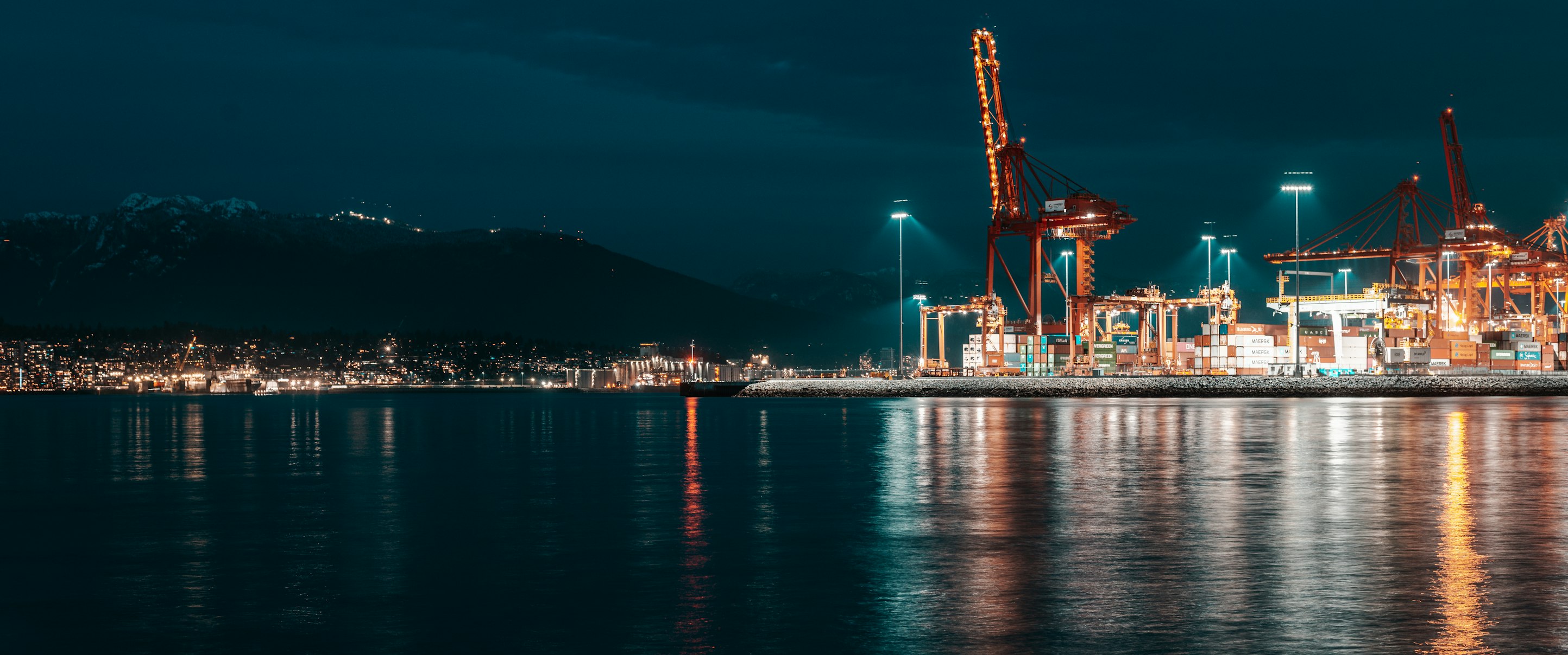 A container port in Vancouver at night: orange cranes and stacked containers reflected in dark harbour water, with snow-dusted mountains and city lights beyond.