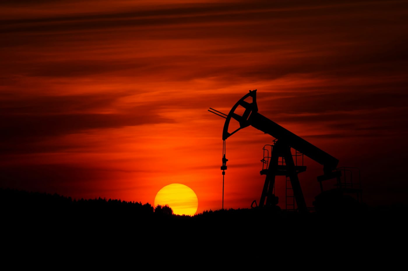 An oil pump jack silhouetted against a deep red sky, with the sun low on the horizon.