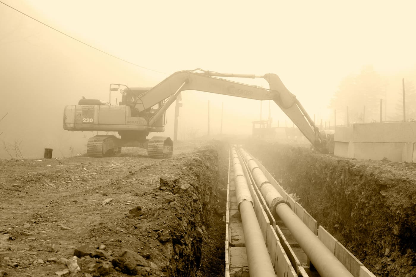 An excavator beside a trench holding large pipes, in morning haze.