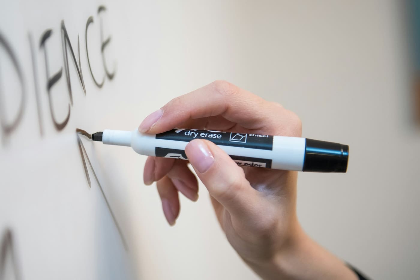 A hand holding a black dry-erase marker writes on a whiteboard.